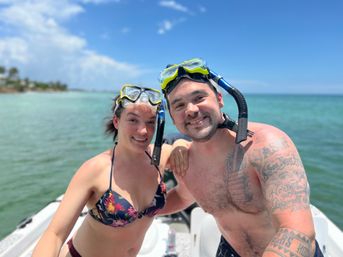 Smiling snorkelers posing on a small boat with bright snorkel masks, turquoise sea and a sunny tropical shoreline under a clear blue sky.