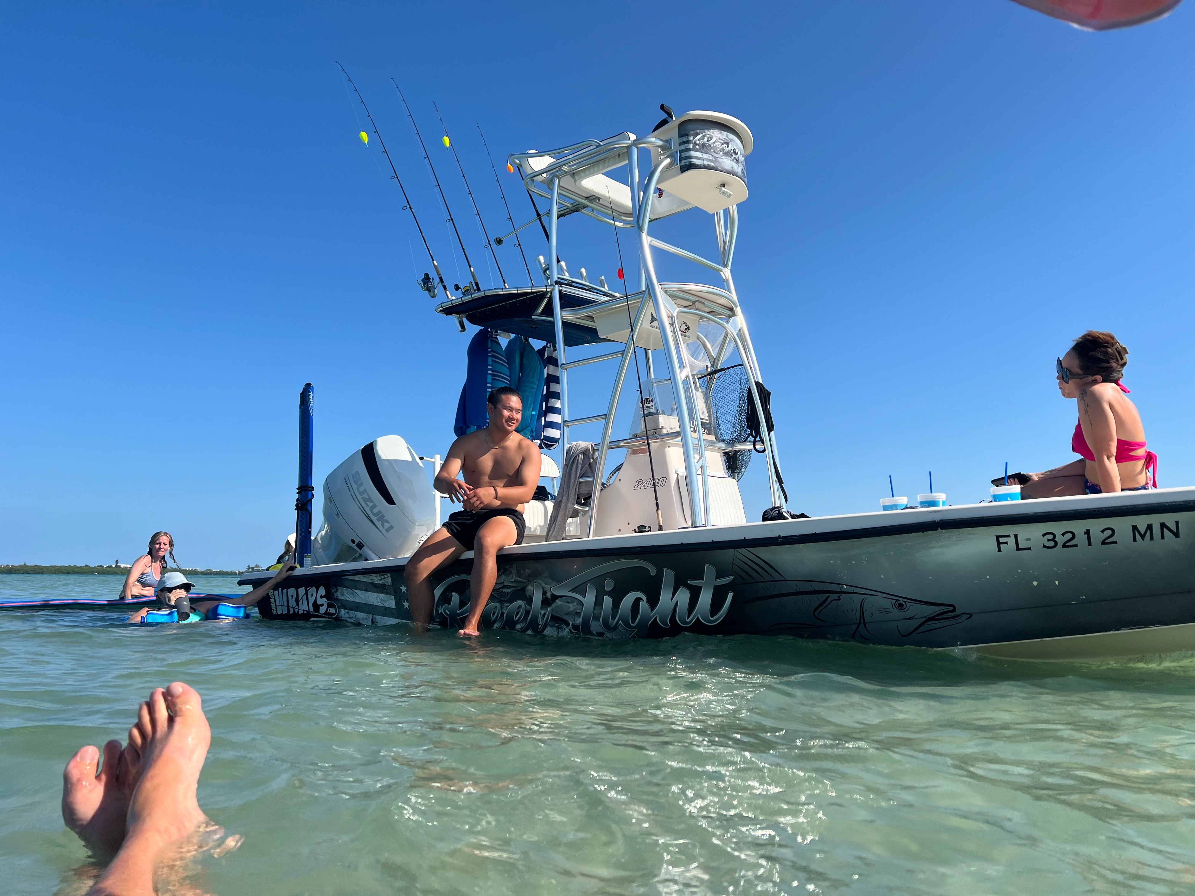 Group relaxing on a center-console boat anchored in shallow, clear turquoise Florida coastal waters under a sunny blue sky, with fishing rods and coolers visible.