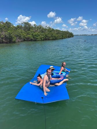 Four people lounging on a bright blue floating mat in a sunny mangrove-lined coastal inlet with turquoise water and puffy clouds — ideal boating and beach-day scene.