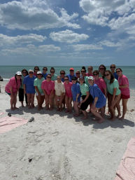 Large group of people in colorful T-shirts posing for a cheerful summer group photo on a sunny sandy beach with turquoise ocean and blue sky dotted with clouds.