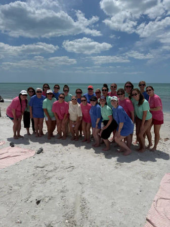 Large group of people in colorful T-shirts posing for a cheerful summer group photo on a sunny sandy beach with turquoise ocean and blue sky dotted with clouds.