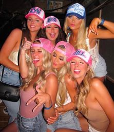 Six women at a lively bar wearing pink and blue personalized trucker hats, posing playfully for a birthday or girls' night out.