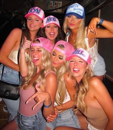 Six women at a lively bar wearing pink and blue personalized trucker hats, posing playfully for a birthday or girls' night out.