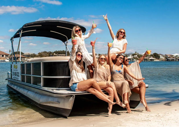 Six women raising colorful cocktails aboard a pontoon boat pulled ashore on a sunny coastal beach, with waterfront homes and blue sky in the background.