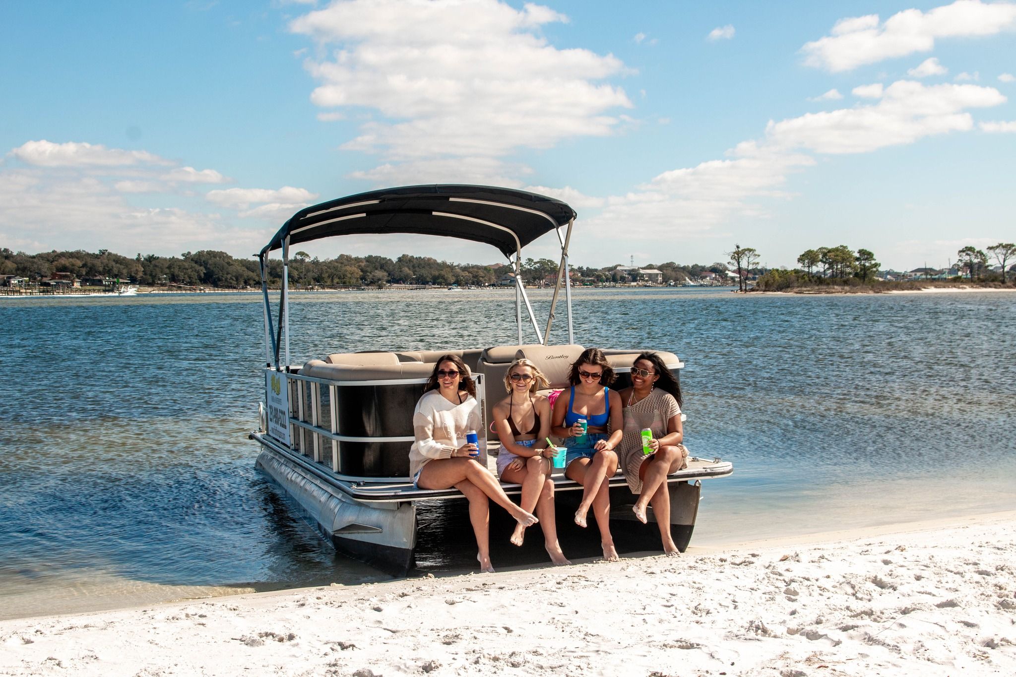 Four friends in swimsuits sitting on the front of a pontoon boat pulled up on a sandy beach, sipping drinks on a sunny day with calm coastal bay waters and blue sky.