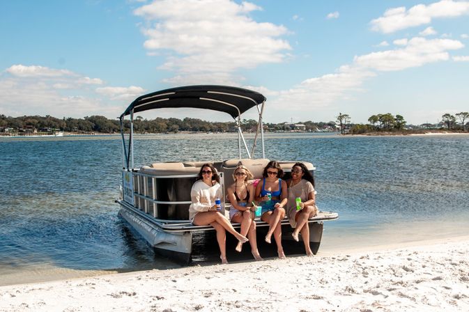Four friends in swimsuits sitting on the front of a pontoon boat pulled up on a sandy beach, sipping drinks on a sunny day with calm coastal bay waters and blue sky.