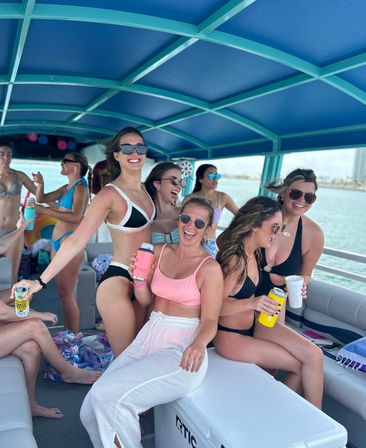 Smiling young women in swimsuits on a covered party boat near a sunny coastline, laughing and holding colorful drink cans and cups under a teal canopy