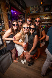 Six women in a cozy wood-paneled bar wearing heart-shaped sunglasses and smiling while posing with drinks — lively nightlife friends’ night out group photo.