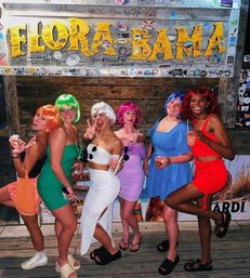 Six people in colorful wigs and summer outfits holding cocktails, posing and smiling in front of a graffiti-covered wooden beach bar sign — upbeat Gulf Coast beach bar nightlife scene.
