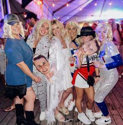 Group of women in blonde wigs and playful costumes posing on a wooden deck under a lit party tent at night, holding oversized face cutouts and drinks — lively outdoor nightlife scene.