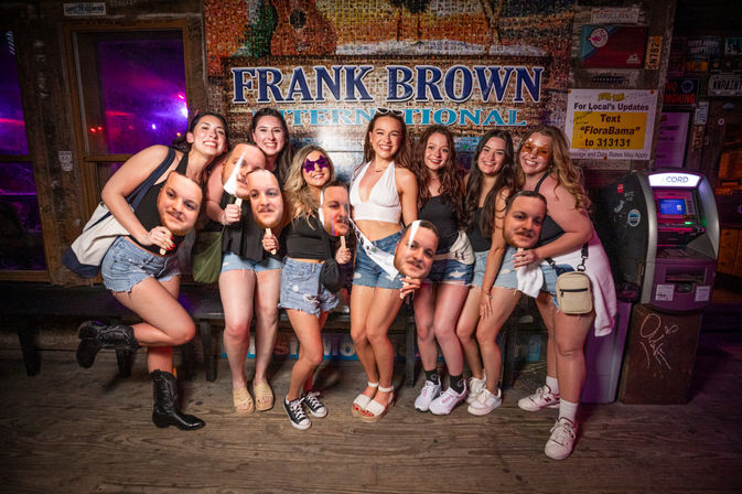 Group of young women in denim shorts posing and laughing at a neon-lit country bar, some holding oversized face cutout masks for a playful bachelorette-style night out.