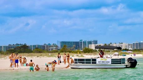 Sunny summer beach scene: a pontoon boat anchored near shore while vacationers swim, sunbathe on white sand and turquoise water with seaside resort buildings in the background.