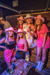 Bachelorette party of five women in pink cowgirl hats and outfits—bride-to-be wearing a sash—smiling and holding drinks in a dim, graffiti-covered bar booth with a carved wooden table.