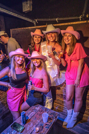 Bachelorette party of five women in pink cowgirl hats and outfits—bride-to-be wearing a sash—smiling and holding drinks in a dim, graffiti-covered bar booth with a carved wooden table.