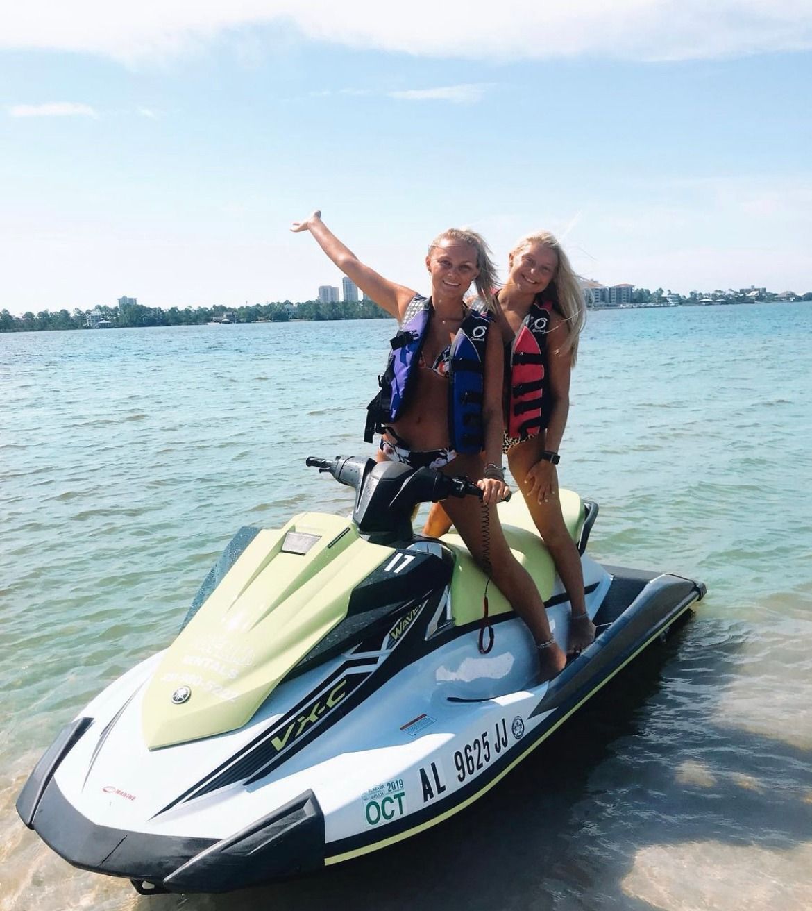 Two smiling women in colorful life jackets posing on a lime-green jet ski in shallow clear blue water off a sunny coastal beach with distant shoreline buildings