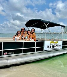 Five friends in swimsuits smiling on a pontoon boat in shallow turquoise water along the Alabama Gulf Coast under a cloudy blue sky.