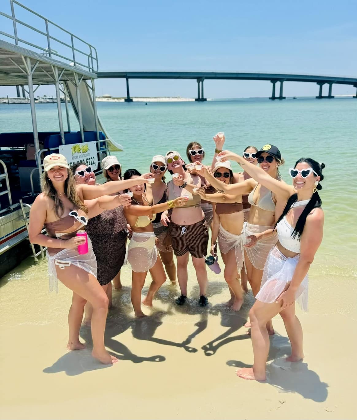 Group of friends in bikinis and heart-shaped sunglasses toasting drinks on a sunny sandy beach beside a pontoon boat, turquoise shallow water and a long coastal bridge in the background.