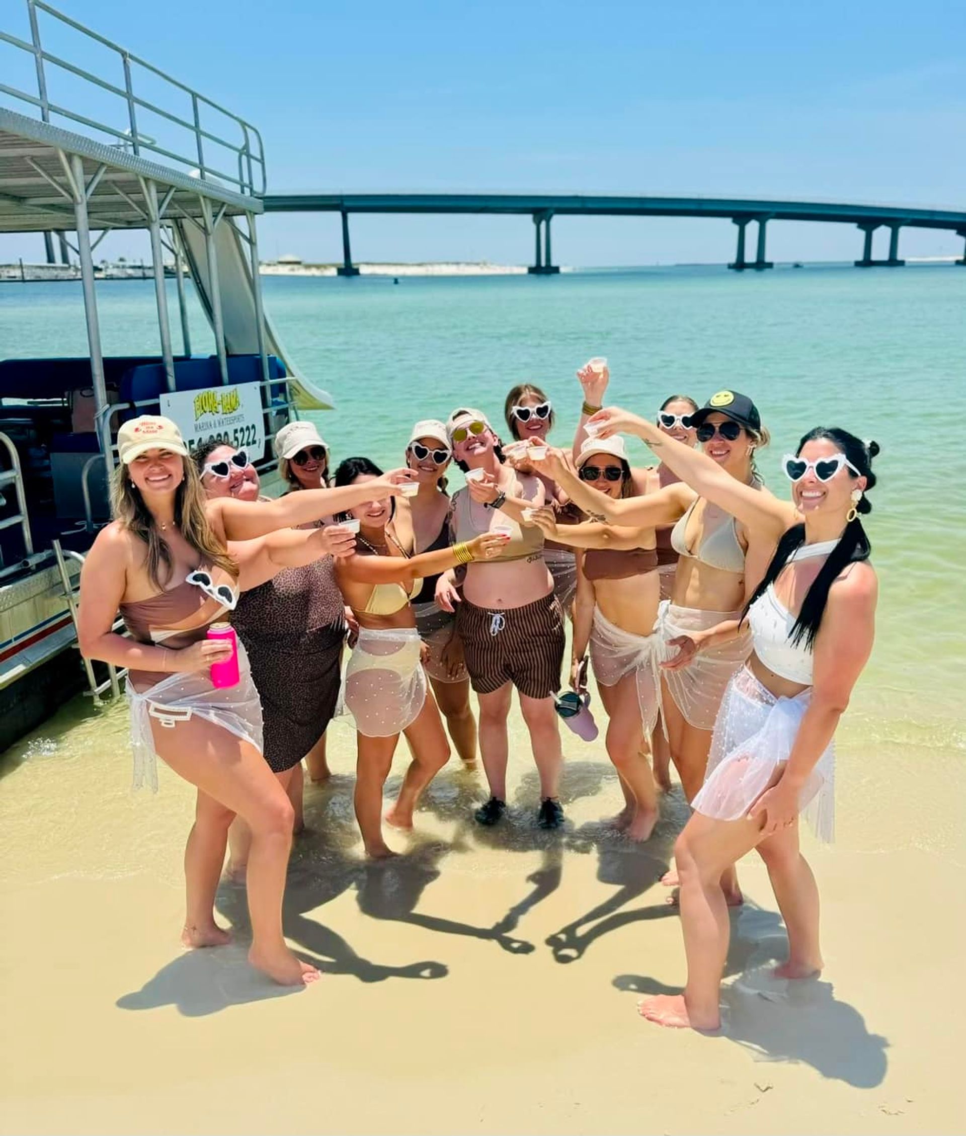 Group of friends in bikinis and heart-shaped sunglasses toasting drinks on a sunny sandy beach beside a pontoon boat, turquoise shallow water and a long coastal bridge in the background.