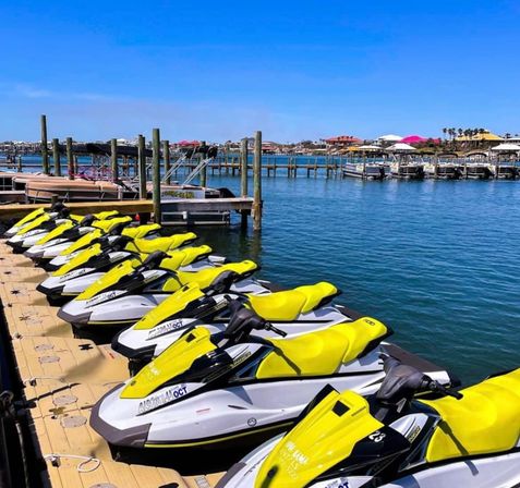 Row of bright yellow jet skis lined up on a floating dock at a sunny coastal marina, calm blue water with colorful beachfront buildings and palm trees in the background.
