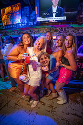 Group of five women at a bachelorette party in a neon-lit coastal bar, wearing bright dresses and a bride sash, posing with drinks and a humorous face cutout amid colorful stickers and TV screens — lively Gulf Coast nightlife vibe.