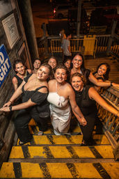 Smiling group of women posing on yellow-striped wooden outdoor stairs at night, celebrating a night out with one woman in white wearing a rosette.
