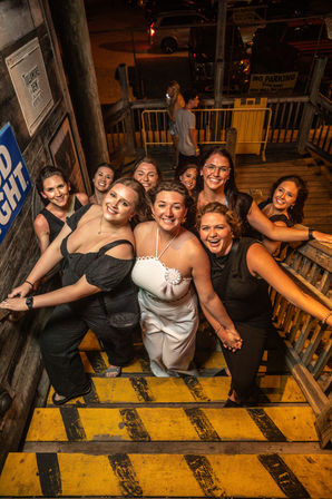 Smiling group of women posing on yellow-striped wooden outdoor stairs at night, celebrating a night out with one woman in white wearing a rosette.