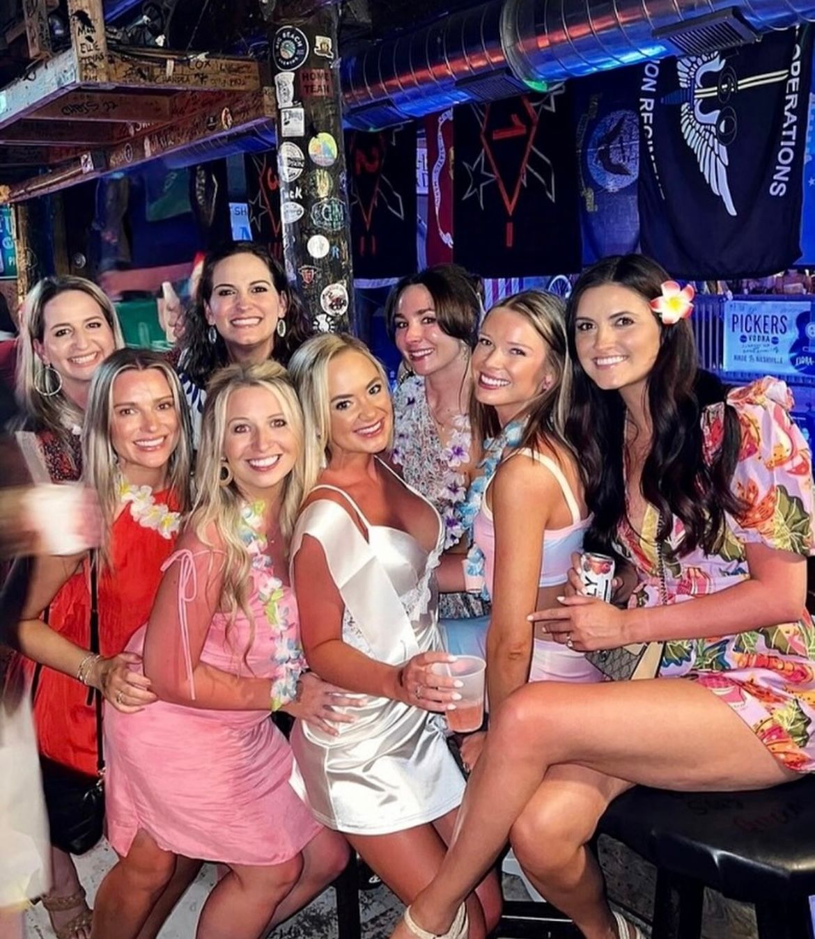 Group of eight smiling women in colorful summer dresses and leis posing at a lively tropical-themed bar with neon lights, a sticker-covered post and hanging flags — girls' night out nightlife photo