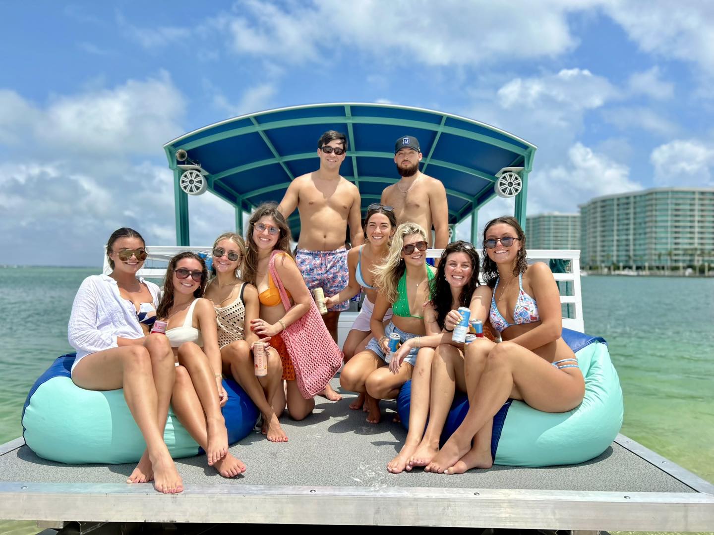 Sun-soaked group of friends in swimsuits smiling on a covered pontoon boat over clear turquoise bay water, holding drinks with beachfront high-rise condos and blue sky in the background.