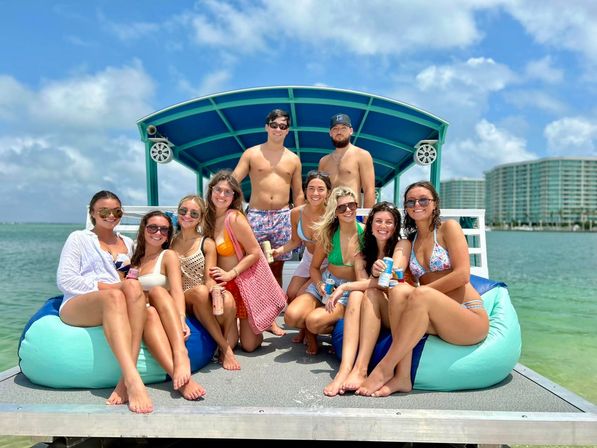 Sun-soaked group of friends in swimsuits smiling on a covered pontoon boat over clear turquoise bay water, holding drinks with beachfront high-rise condos and blue sky in the background.