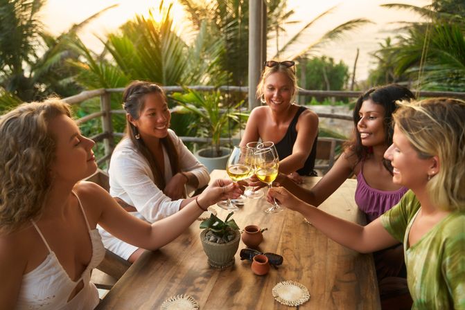 Five friends clinking white-wine glasses around a wooden table on a tropical outdoor terrace at sunset with palm trees in the background.