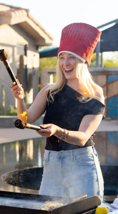 Playful person in a tall red hat holding two flaming torches over a flat-top grill on a sunny backyard poolside patio.