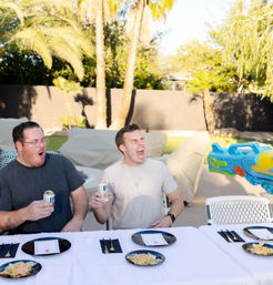 Two men at a sunny backyard table holding canned drinks and reacting as a colorful water gun sprays them, with salad plates and palm trees in the background.
