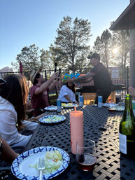 Outdoor patio dinner at sunset with friends laughing as one person hands a colorful water gun across a table of paper plates, drinks, and salad, pine trees and red rock cliffs in the background.