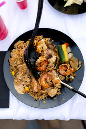 Grilled shrimp and chicken over fried rice with creamy sauce, grilled zucchini and mixed vegetables on a black plate at an outdoor table with red cups