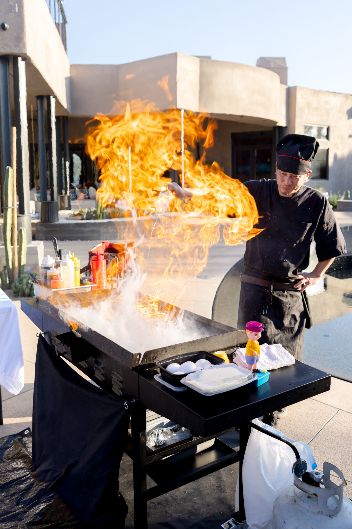 Outdoor chef cooking on a flat-top griddle with dramatic flambé flames on a sunny southwestern-style patio, with condiments, eggs and a propane tank nearby