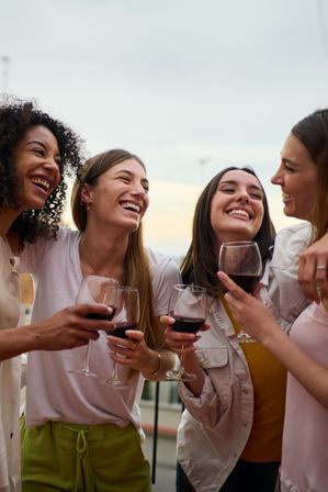 Four women laughing on a rooftop terrace at sunset, toasting glasses of red wine during a casual friends' night