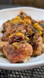 Close-up of roasted chicken drumsticks topped with caramelized golden raisins, fresh herbs and a glossy savory glaze in a white bowl on a metal patio table