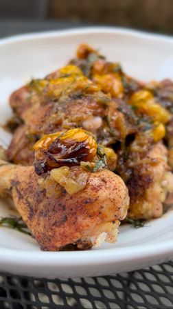 Close-up of roasted chicken drumsticks topped with caramelized golden raisins, fresh herbs and a glossy savory glaze in a white bowl on a metal patio table
