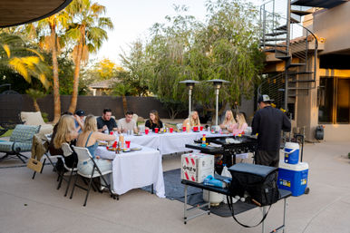 Friends enjoying an outdoor dinner party on a modern backyard patio with palm trees, long white-tablecloth tables and red cups, a chef at a portable grill, patio heaters and a spiral staircase.