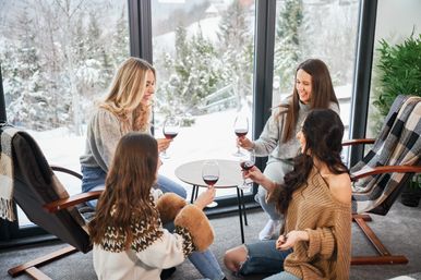 Four friends in cozy sweaters toast red wine by floor-to-ceiling windows overlooking a snowy forest — relaxed winter cabin gathering.