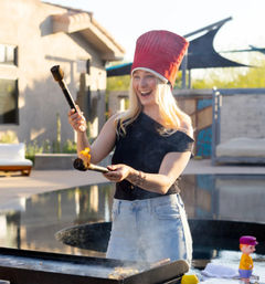 Person wearing a tall red chef hat laughing while holding two flaming skewers over a griddle on a sunny backyard pool patio, casual outdoor cooking scene.