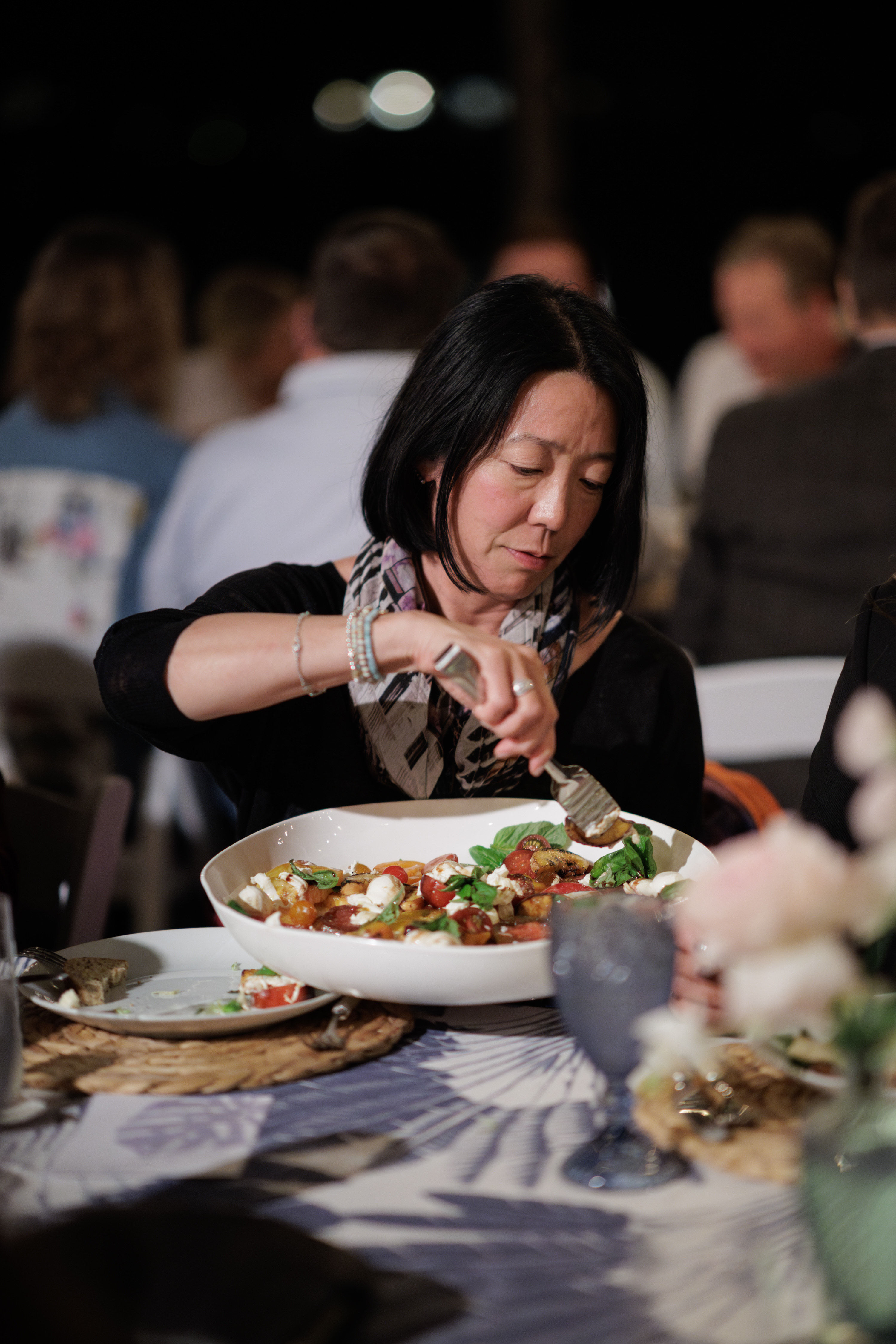 Woman serving a tomato-and-mozzarella salad from a large white bowl at a lively dinner party table with blurred guests