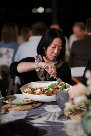 Woman serving a tomato-and-mozzarella salad from a large white bowl at a lively dinner party table with blurred guests