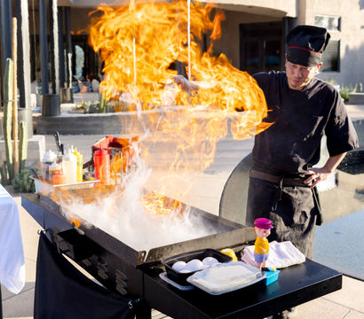 Outdoor chef in black uniform flambés on a smoking griddle at a sunny desert poolside patio — towering orange flame, condiments and eggs on the cart with a propane tank nearby.