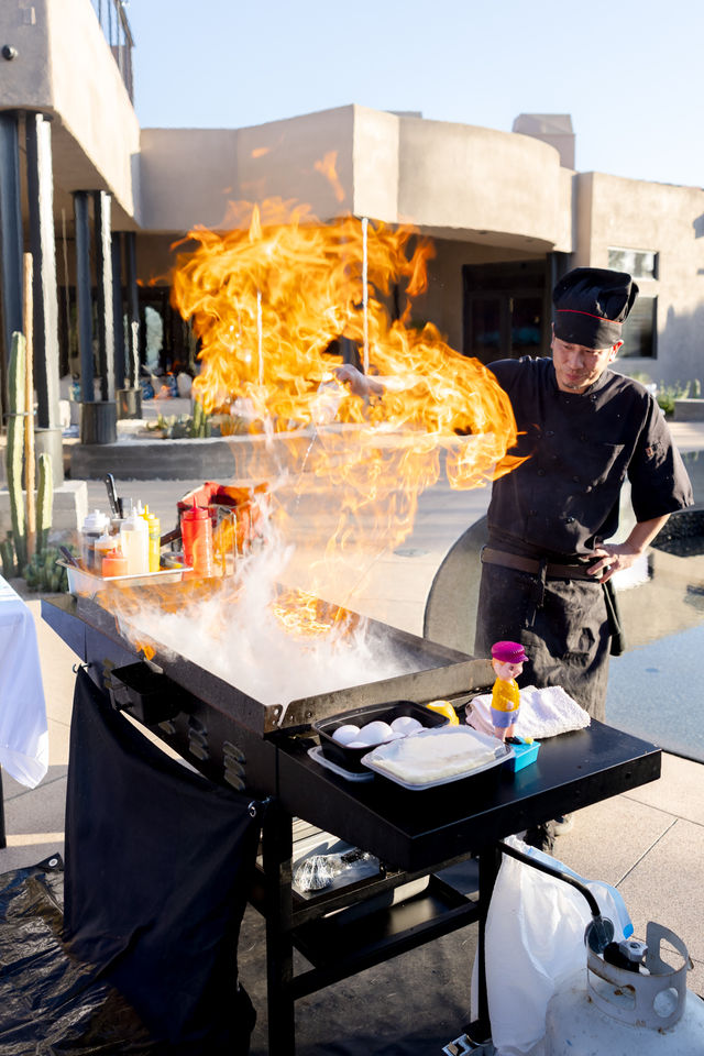 Outdoor chef in black uniform flambés on a smoking griddle at a sunny desert poolside patio — towering orange flame, condiments and eggs on the cart with a propane tank nearby.