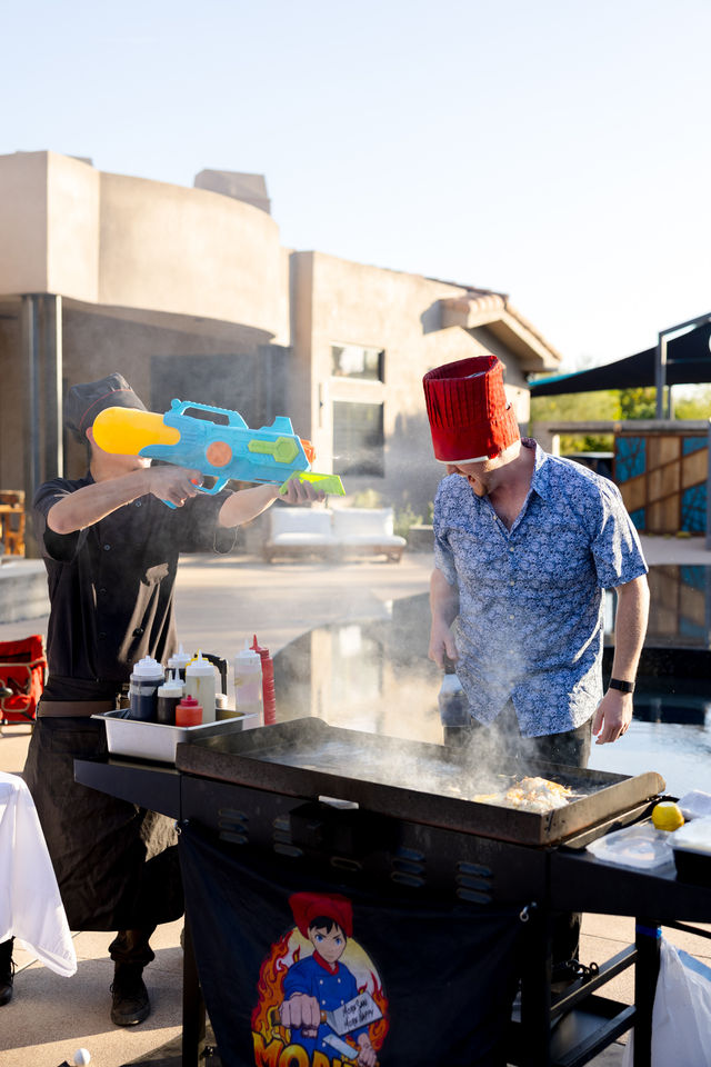 Playful poolside cookout: two adults at a flat-top griddle beside a modern backyard pool, one wearing a red hat flipping food amid steam while the other fires a colorful toy water gun, condiments and spatula nearby.