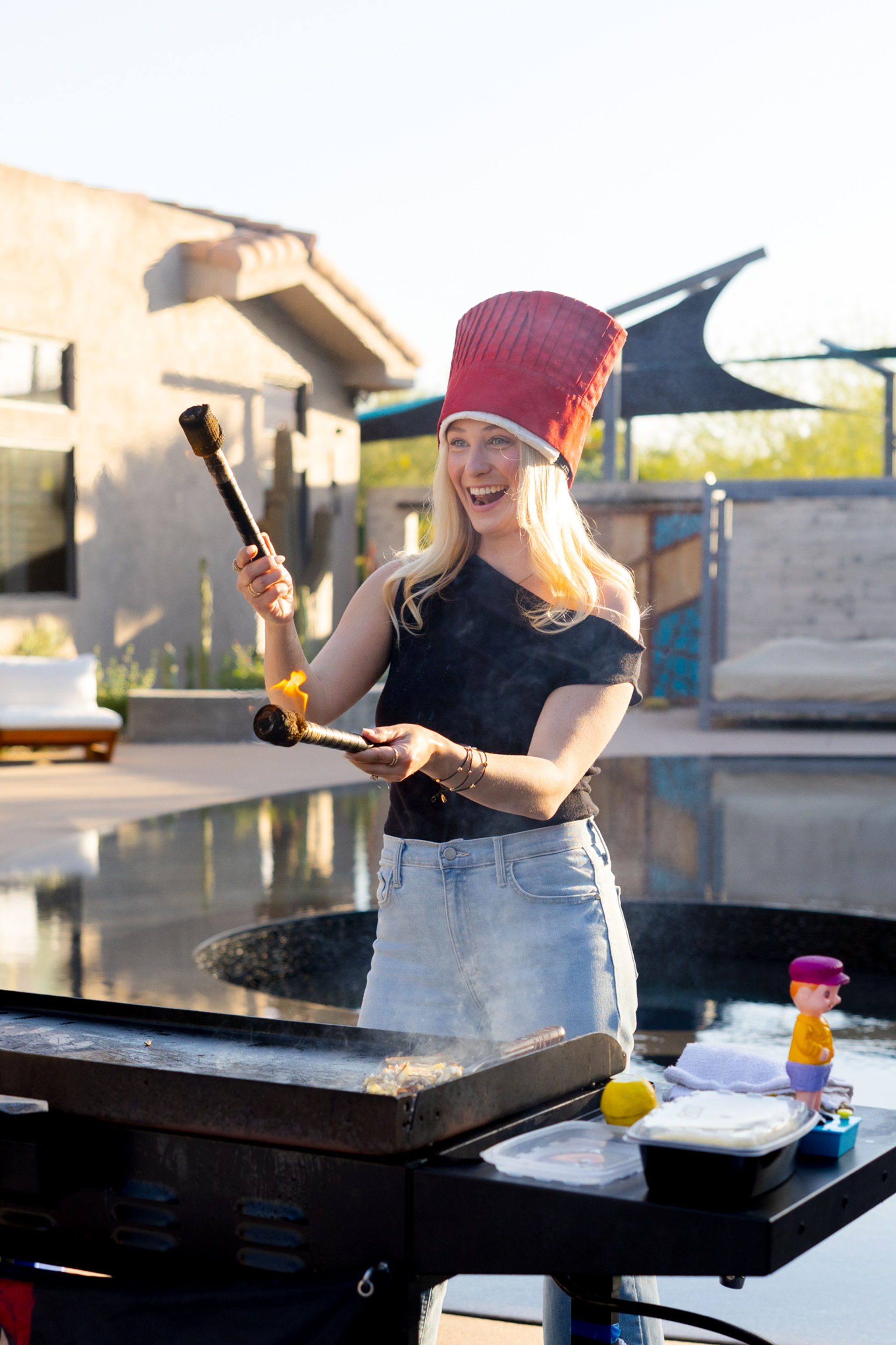 Smiling person in a tall red chef hat juggling flaming torches over a griddle on a sunny backyard patio beside a pool.