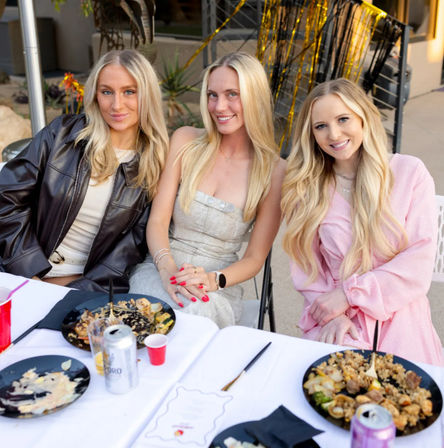 Three smiling blonde friends in stylish outfits seated at an outdoor patio dinner table with plates of food, drinks, and gold party streamers in the background.