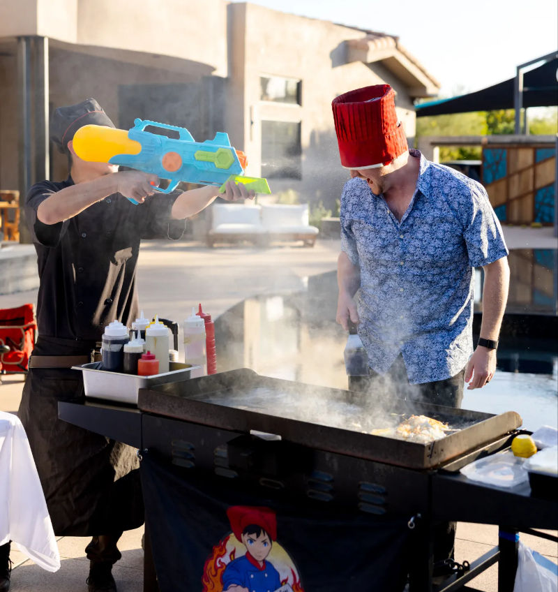 Outdoor patio cookout by a pool: person wearing a red bucket hat tending a steaming flat-top griddle as another playfully sprays them with a large colorful water gun, condiment bottles lined on the grill station.