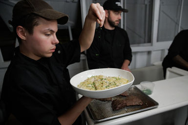 Cook in a commercial kitchen sprinkling fresh herbs over a bowl of creamy risotto, with grilled steak on a wire rack and prep containers on the counter